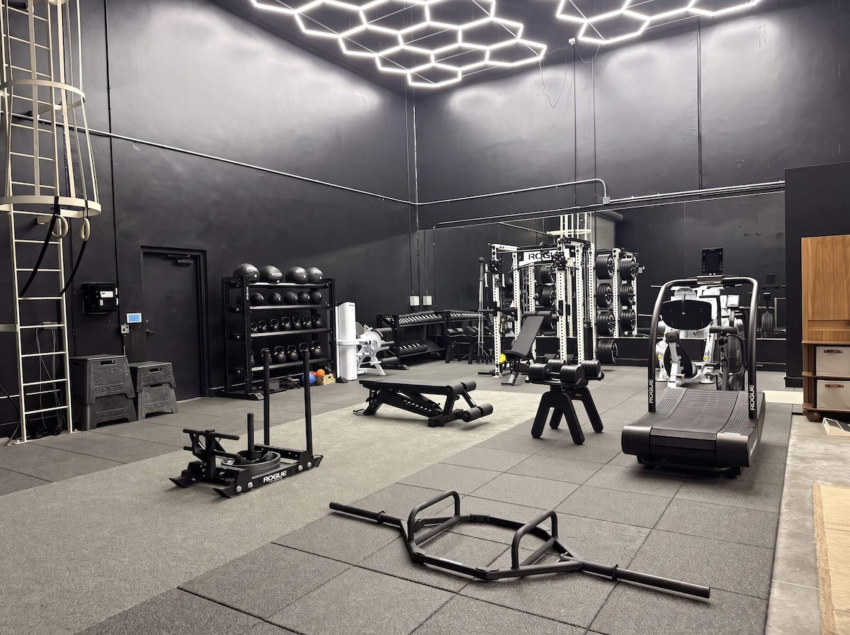 Blacked-out warehouse gym floor with sled, benches, and treadmill under hexagon LED lighting